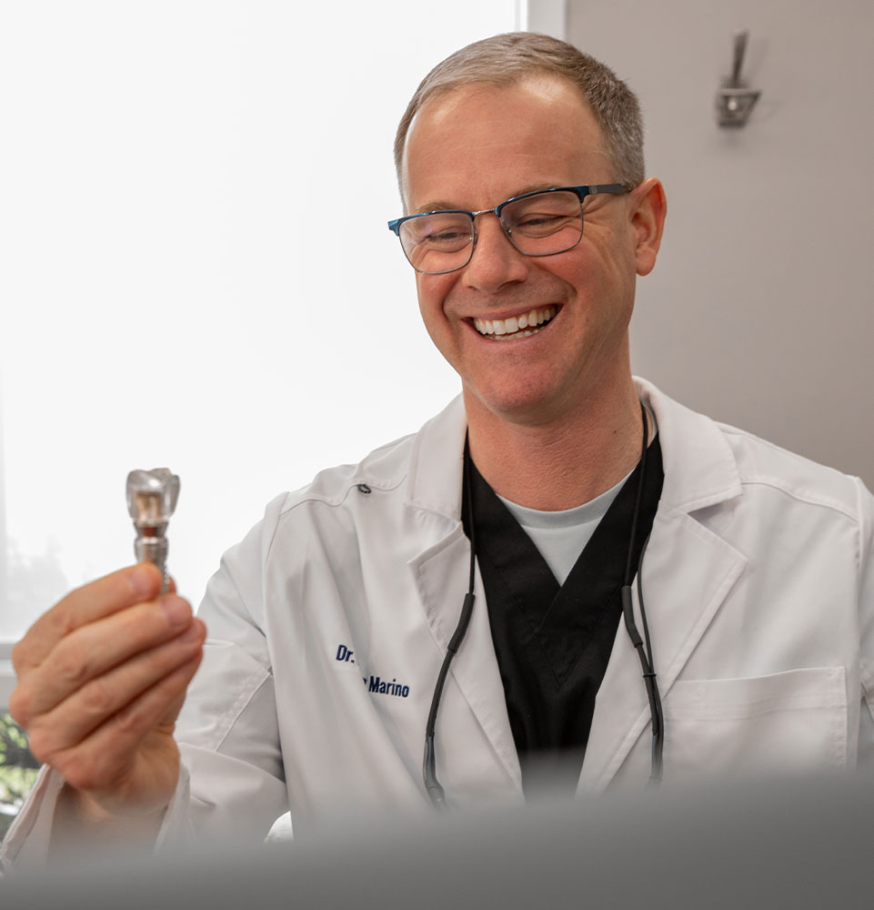 Dr. Brian Marino smiling as he examines a dental implant abutment at The Center for Dentistry of Vernon in Vernon Township, New Jersey